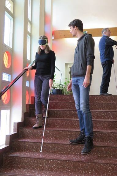 Selbsterfahrung mit Taststock und Augenbinde. Eine Schülerin geht mit Augenbinde und Langstock die Stufen runter. Ein Schüler beobachtet sie dabei und hilft, wenn nötig.