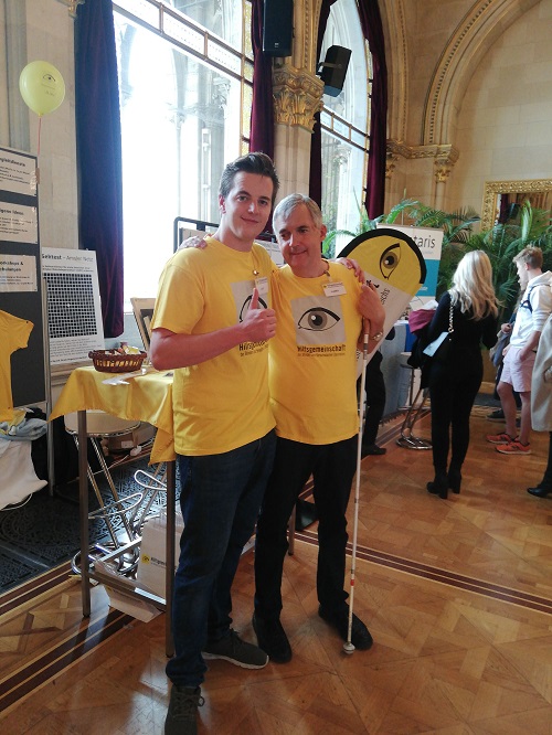 Maximilian Reutterer mit Team-Mitglied Harry Rother bei der Freiwilligenmesse im Wiener Rathaus Zwei Männer in gelben T-Shirts stehen nebeneinander in einem Saal auf einer Messe. Der ältere Mann hat einen weißen Stock.