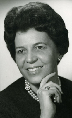 Augustine Mayer Schwarz-weiß Foto einer lächelnden Dame mittleren Alters, die ihre Hand ans Kinn hält und nicht direkt in die Kamera blickt