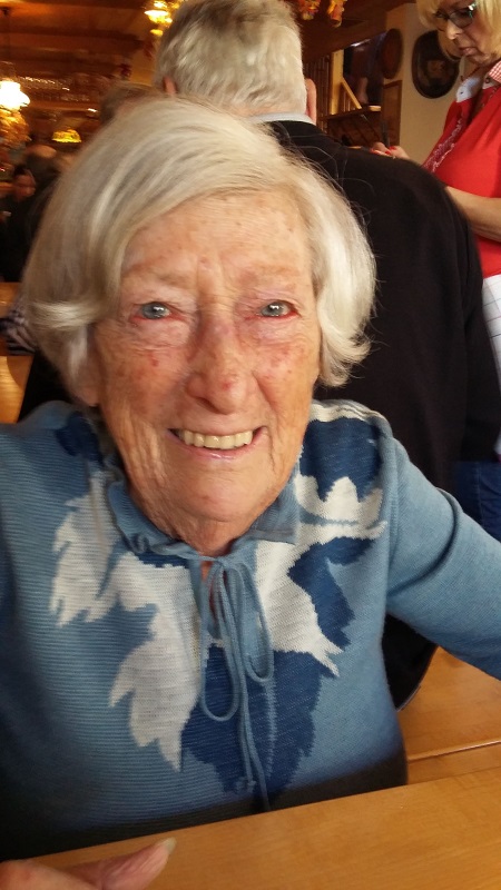 Erika Feichtinger Eine ältere Dame lächelt fröhlich in die Kamera, sie sitzt an einem Tisch, hinter ihr sieht man Menschen