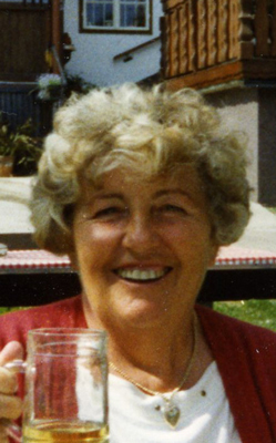 Wilhelmine Frischauf Eine ältere Dame mit Dauerwelle hält ein Glas in der Hand und lächelt fröhlich in die Kamera, hinter ihr sieht man einen Teil eines alten Bauernhauses