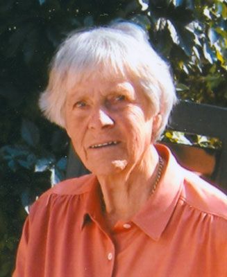 Janna Prast Eine ältere Dame sitzt vor einer grünen Hecke und blickt leicht lächelnd in die Kamera