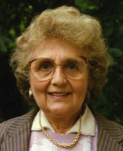 Gertrude Lesak Eine ältere Dame mit großer Brille steht vor einer grünen Hecke und lächelt freundlich in die Kamera