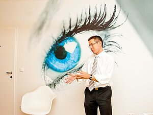 Priv. Doz. Dr. Erdem Ergun, Copyright: Erdem Ergun Ein Mann mit Brille steht gestikulierend vor einer Wand auf der ein großes blaues Auge zu sehen ist