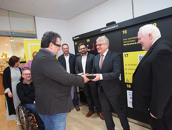 Mag. Klaus Höckner übergibt dem Bürgermeister Klaus Peter Luger (SPÖ) eine Linzertrorte. Mann übergibt Torte an anderen Herren vor Paketbox