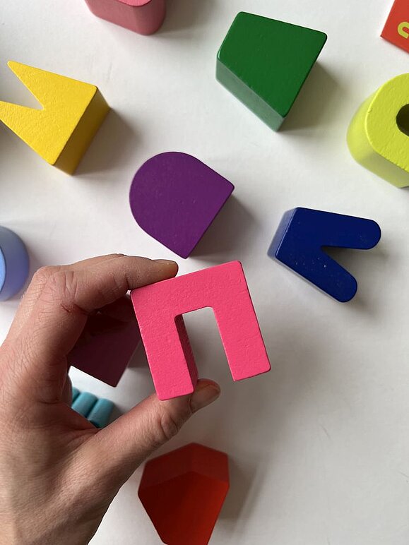 talking hands hat mit tilo ein Tastspiel aus Holz entwickelt, das wie Memory funktioniert. Hand hebt rosa umgekehrtes "U" aus Holz von weißer Oberfläche, dahinter liegen weitere bunte Formen