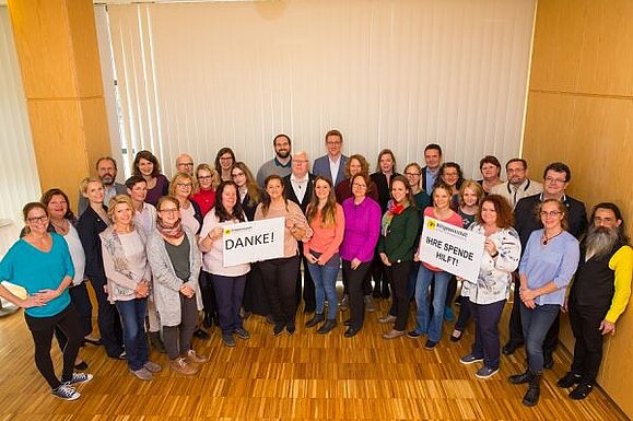 Die Mitarbeiter:innen der Hilfsgemeinschaft stehen in einem Halbkreis in einem Raum nebeneinander. Zwei Schilder werden hoch gehalten. Eines mit "Danke" Aufschrift, ein weiteres mit "Ihre Spende hilft!". 
