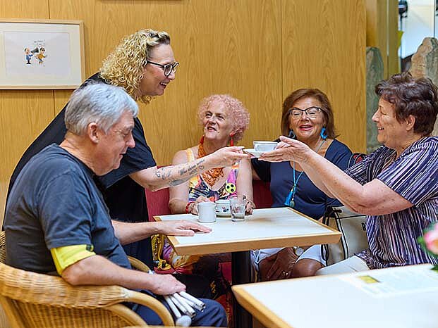 Ein Mann und drei Frauen sitzen bei einem Tisch. Eine weitere Frau steht und gibt der sitzenden Frau einen Kaffee. Am Tisch stehen Kaffeetassen und an einem Tisch im Vordergrund stehen rosa Blumen..