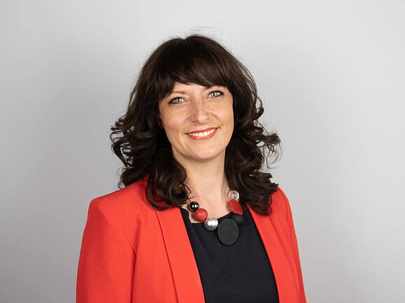 Frau mit dunkelbraunen gelockten Haaren und rotem Blazer lächelt in die Kamera