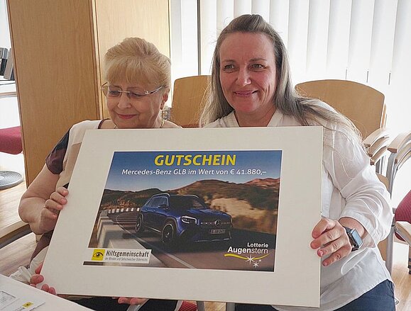 Die Gewinnerin des Hauptpreises spendet seit Jahrzehnten für die Hilfsgemeinschaft der Blinden und Sehschwachen Österreichs. Ältere und jüngere Frau halten Gutschein mit Bild von Auto