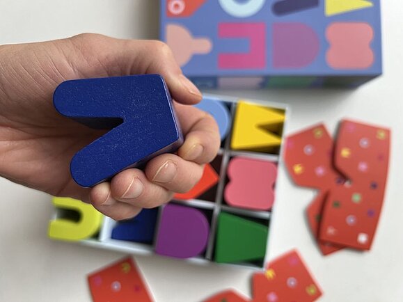 Die Figuren von tilo sind aus Holz. Hand hält blaues "V" aus Holz, dahinter sieht man Spieleschachtel mit bunten Formen