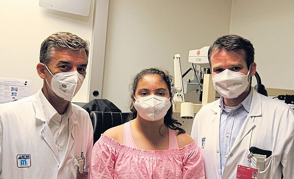 Netzhautchirurg Michael Georgopoulos (li.), Gian, Markus Ritter. Zwei Ärzte und Mädchen mit rosa Shirt schauen in die Kamera.