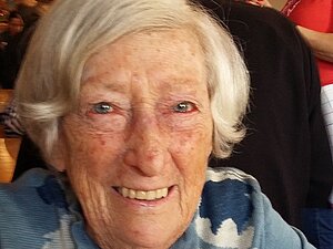 Erika Feichtinger Eine ältere Dame lächelt fröhlich in die Kamera, sie sitzt an einem Tisch, hinter ihr sieht man Menschen