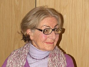 Elfriede Trulp Eine ältere Dame mit Brille und Schal sitzt lächelnd an einem Tisch