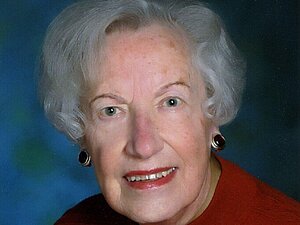 Edith Simanek Eine ältere Dame mit grauer Dauerwelle, großen Ohrringen und rotem Lippenstift lächelt in die Kamera.