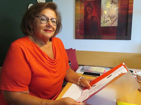 Helene ist über ihre Mutter zur Hilfsgemeinschaft gekommen und hat hier neue Freunde und viel Freude in der Singgruppe gefunden. Helene, braunhaarige Frau mit orangenem Pullover und Brille sitzt an einem Tisch mit einem Folder in der Hand und lächelt.