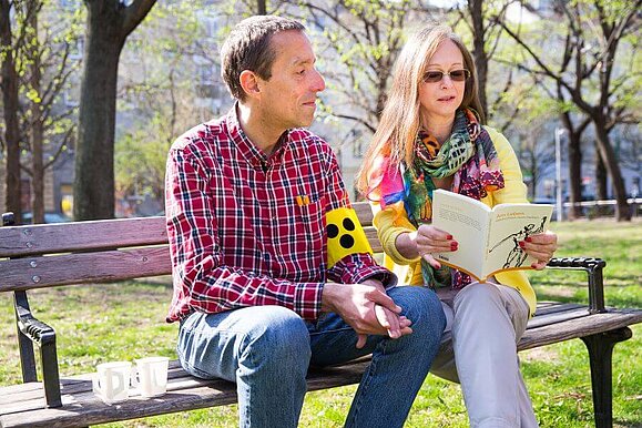 Beim Begleitdienst stehen Freizeitaktivitäten wie Plaudern, Spazieren und Vorlesen im Mittelpunkt Zwei Personen sitzen auf einer Bank. Eine Person trägt eine Armschleife, die andere liest etwas vor.