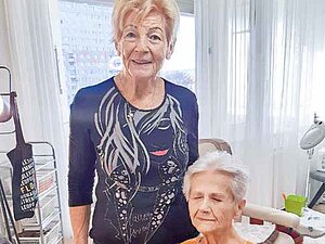 Zwei ältere Frauen, eine sitzt, in einem Pflegezimmer.