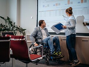 Mann mit Rollstuhl und stehende Frau im Büro schauen sich Zahlen auf einer Leinwand an
