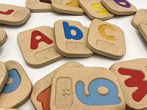 Eine Sammlung von Holzklötzen, auf denen farbige Buchstaben angebracht sind. Darunter steht der jeweilige Buichstabe in Braille Schrift. Jeder Klotz ist rechteckig mit abgerundeten Kanten. 