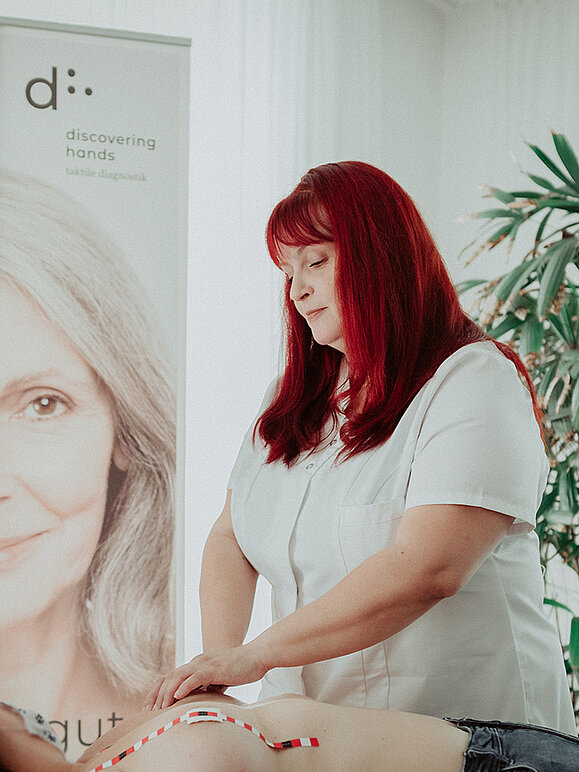 Eine Frau mit knallroten Haaren trägt weißen Kittel und tastet die Brust einer Frau ab, die vor ihr liegt. Im Hintergrund steht ein Banner mit dem Logo von discovering hands und dem Porträt einer grauhaarigen lächelnden Frau.
