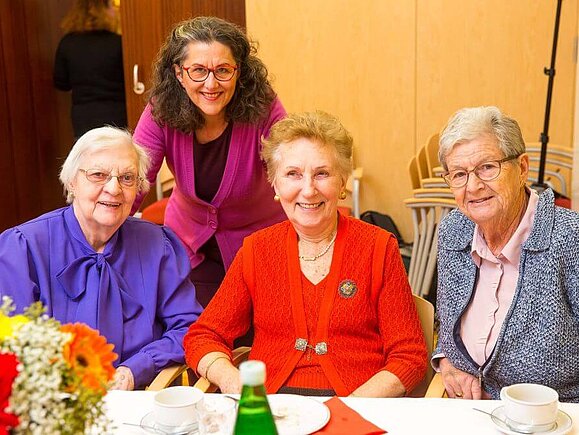 Frau J. sitzt bei einem Testatoren-Treffen mit zwei weiteren Damen an einem Tisch, im Hintergrund ist die Mitarbeiterin der Hilfsgemeinschaft, Frau Portner-Frisch zu sehen.