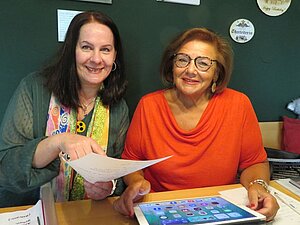 Die Freundinnen Gaby S. und Helene haben immer viel Freude beim gemeinsamen Vorbereiten der Auftriffte der Singgruppe. Zwei Frauen sitzen am Tischen und haben Unterlagen in der Hand, sie lächeln freundlich in die Kamera.