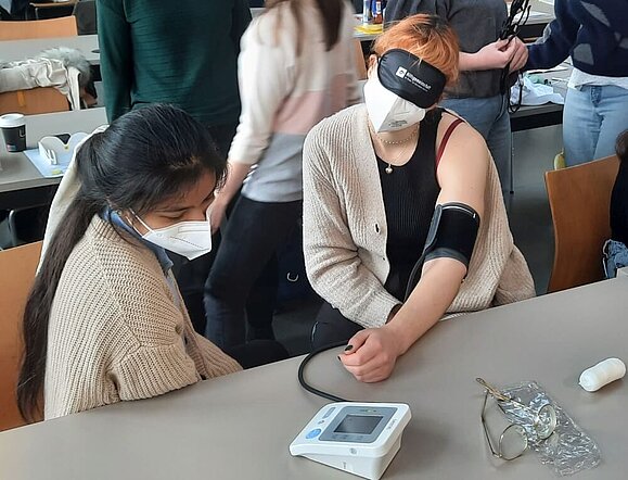 reiter_Schulungen_Pflegeschulung junge Frau misst Blutdruck einer anderen jungen Frau mit Augenbinde