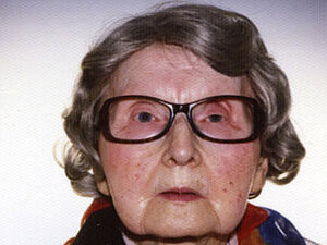 Lydia Digles Eine ältere Dame mit großer Brille blickt ernst in die Kamera