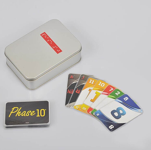 Phase 10 Spiel mit Braille-Schrift Eine Metallbox mit Braille-Schrift, eine Karte mit dem Phase 10 Logo und mehrere farbige Zahlenkarten mit Prägung liegen am Tisch.
