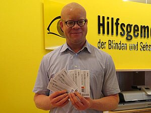 Wir helfen in der Not! Ein Mann mit Brille und Glatze hält 5 REWE Einkaufsgutscheine in den Händen. Hinter ihm sieht man das Hilfsgemeinschaft-Logo.