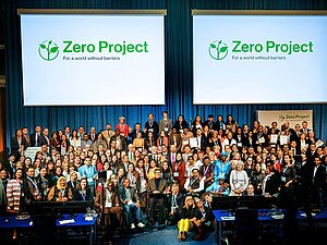 Viele Menschen stehen auf einer Bühne nebeneinander. Darüber sieht man 2x das Zero Project Logo auf einer Leinwand.