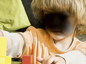 Die AMD-typischen dunklen Flecken im zentralen Gesichtsfeld bereiten auch Probleme beim Erkennen von Gesichtern. Ein kleines Kind mit einem großen, schwarzen Fleck vor dem Gesicht, spielt mit Holzbausteinen. Der Fleck symbolisiert die Sicht eines Menschen, der an AMD, Alterbedingter Makula-Degeneration leidet.