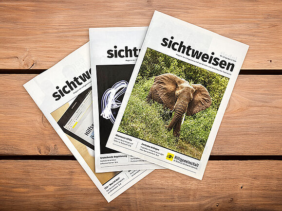 Auf einem Holztisch liegen aufgefächert drei Ausgaben der Sichtweisen. Das Titelbild auf der Obersten zeigt einen Elefanten, der aus Gebüsch herausragt.