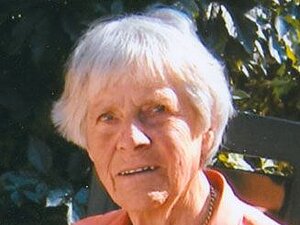 Janna Prast Eine ältere Dame sitzt vor einer grünen Hecke und blickt leicht lächelnd in die Kamera