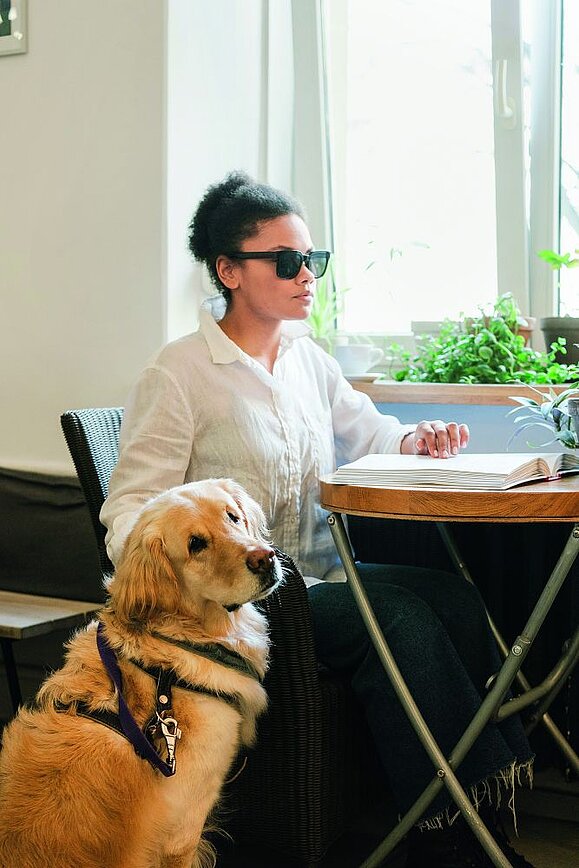 Blinde Frau sitzt an Tisch und liest Braille mit einer Hand und andere Hand auf Hund