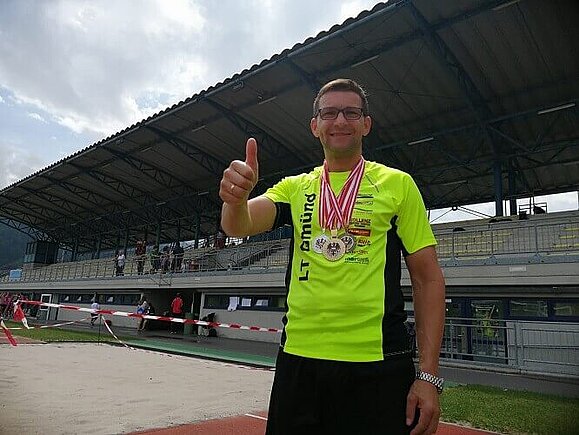 Franz Haider bei der Österreichischen Meisterschaft für Blinde und Sehbehinderte in Innsbruck, Copyright: Franz Haider Ein Mann steht mit neongelben T-Shirt und drei Medaillen um den Hals in einem Stadion. Er hält den erhobenen Daumen in die Kamera.