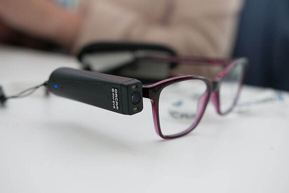 Neueste Version der OrCam. Rote Brille, am rechten Bügel OrCam Gerät befestigt.