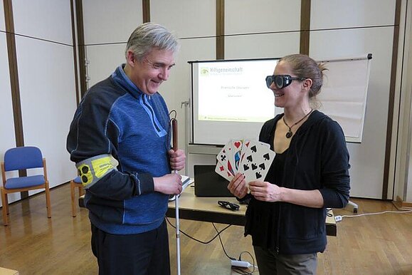 Petra Wrba und Harald Rother beim Workshop mit den Freiwilligen. Petra Wrba hält große Tarockkarten in den Händen, sie trägt dabei eine Simulationsbrille. Harald Rother steht mit Taststock und Armbinde daneben.