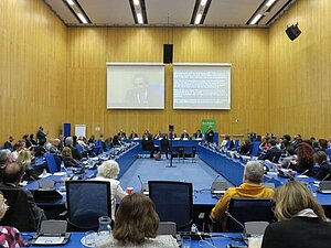 Großer Saal mit Podium und vielen Besuchern. Hinter dem Podium hängen 2 Bildschirme für die Übertragung der Vorträge.
