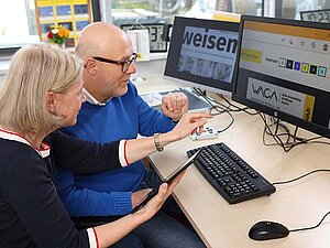 Eine Frau und ein Mann sitzen vor zwei großen Bildschirmen, auf denen in stark vergrößerter Version Websiten dargestellt sind. Die Fraut hält auch ein Tablet in der Hand.