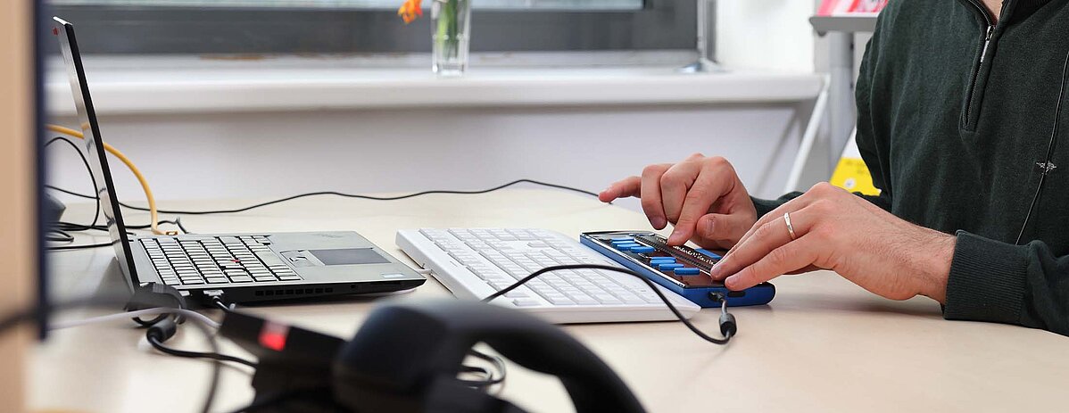 Mann mit Sehbehinderung sitzt mit Headset vor einem Computer mit Braillezeile und arbeitet.