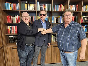 Drei Männer schauen in die Kamera und halten die Hände aufeinander. Elmar Fürst (Vorstandsvorsitzender der Hilfsgemeinschaft), Christian Zehetgruber (Geschäftsführer von VIDEBIS) und Klaus Höckner (Stellvertretender Vorstandsvorsitzender der Hilfsgemeinschaft)