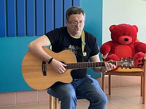 „Musik ist für mich so wichtig wie die Luft zum atmen“, sagt Ruslan. Ruslan, ein hochgradig sehbehinderter Musikliebhaber aus der Ukraine, der derzeit in der Waldpension lebt, sitzt auf einem Schemel und spielt Gitarre vor einer blauen Wand. Neben ihm sitzt ein roter Teddybär auf einem Sessel.