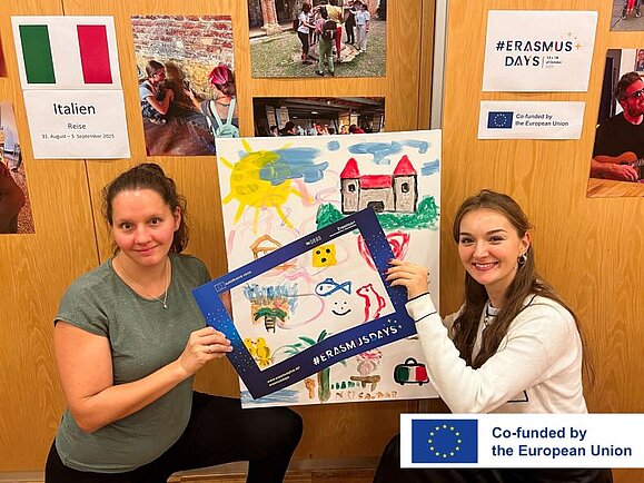 Eröffnung der Fotoausstellung Zwei junge Frauen halten einen Bilderrahmen, im Hintergrund sieht man Fotos