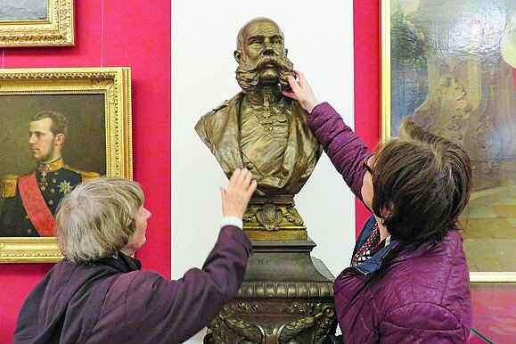 Kulturblick für alle_Hilfsgemeinschaft Zwei ältere Frauen von hinten ertasten eine goldene Büste eines Kaisers