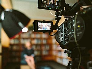 TV-Kamera ist auf eine Person gerichtet, die vor einer Bücherwand sitzt, die Person und die Wand sind unscharf im Hintergrund zu sehen. 