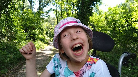Die mehrfach behinderte Anna sitzt in ihrem Rollstuhl im Grünen und freut sich mit einem breiten, ansteckendem Lächeln