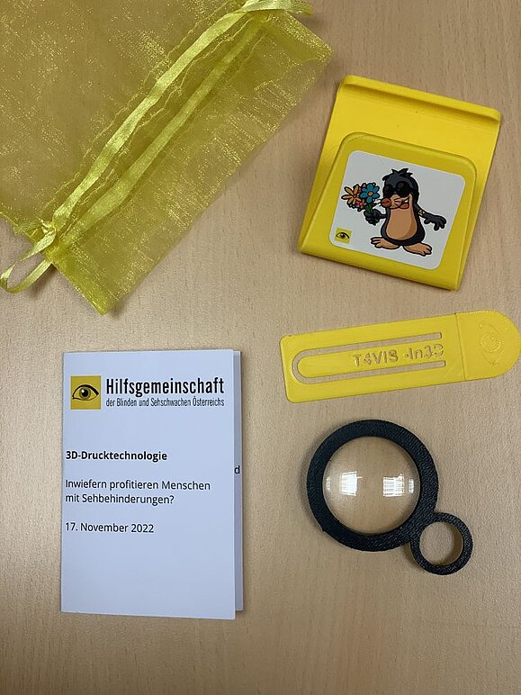 3D-Drucker-Hilfsmittel-Event_Goodiebag.jpg Gelbes Täschchen, schwarze kleine Lupe, gelber Handystehhalter mit Maulwurfgrafik, gelbes Lesezeichen und Papierkärtchen mit Logo der Hilfsgemeinschaft