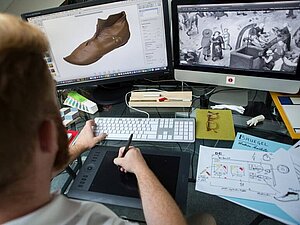 Mann sitzt vor 2 Bildschirmen und Tastatur, in seiner Hand ein Stift für den Computer, Copyright: Tactile Studio Paris